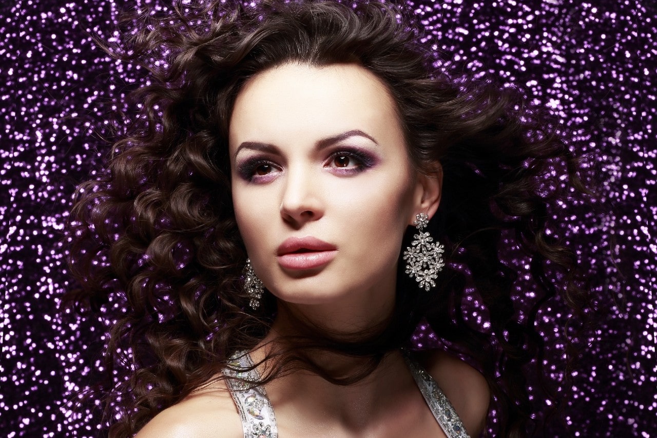 a model wearing diamond earrings stands in front of a purple glittery backdrop.