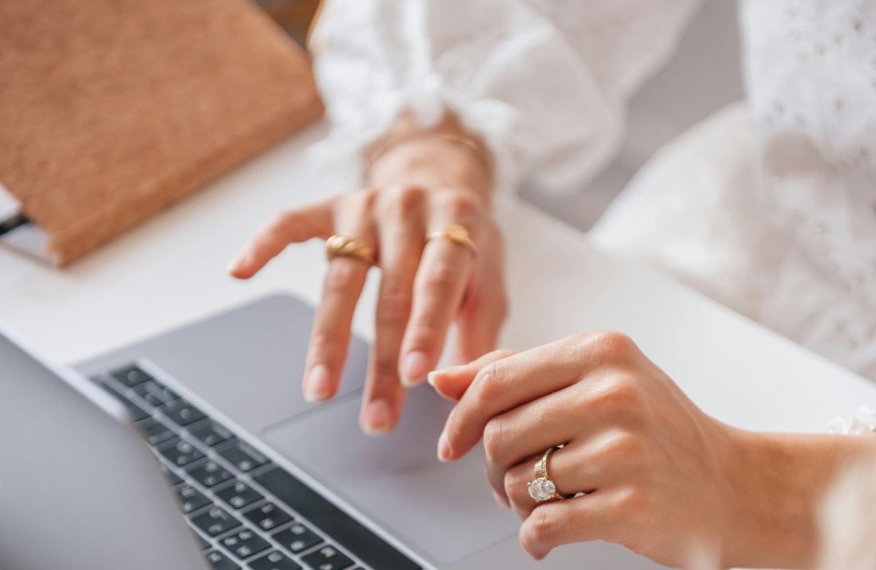 A close-up of a woman’s hands typing on a laptop keyboard, adorned with gold rings and a large diamond engagement ring.