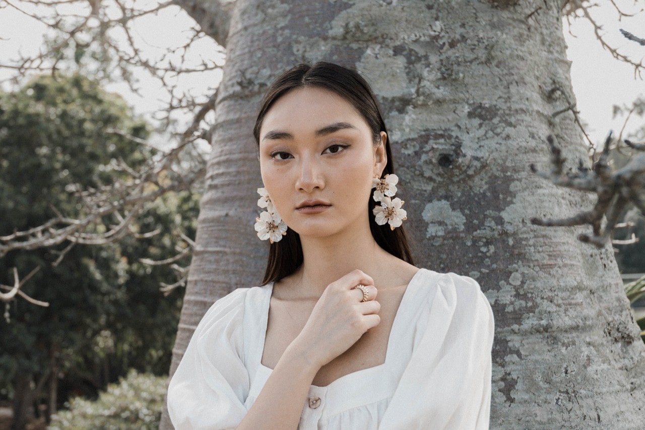 a woman standing in front of a tree in the springtime, wearing a white dress and floral earrings