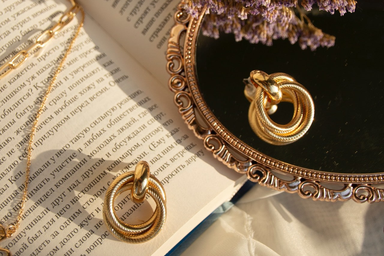 A pair of gold earrings and necklace sit on an open book with a vintage mirror and lavender.