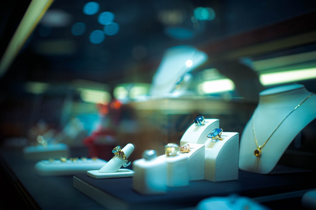 A luxurious jewelry display features an array of bold gemstone rings and a gold necklace, set against a moody, high-end boutique background.