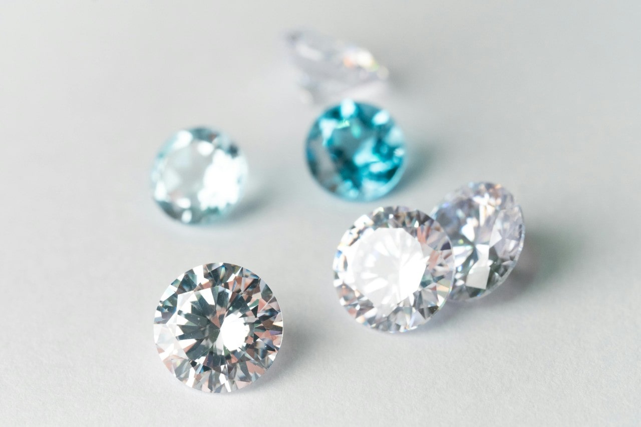 Scattered gemstones on a white surface, featuring four sparkling clear diamonds and two shimmering blue gems, reflecting light.