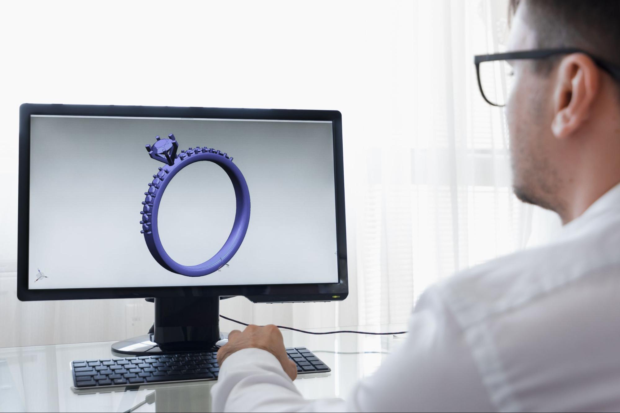A jeweler wearing glasses finalizes a 3D ring design on a computer screen.