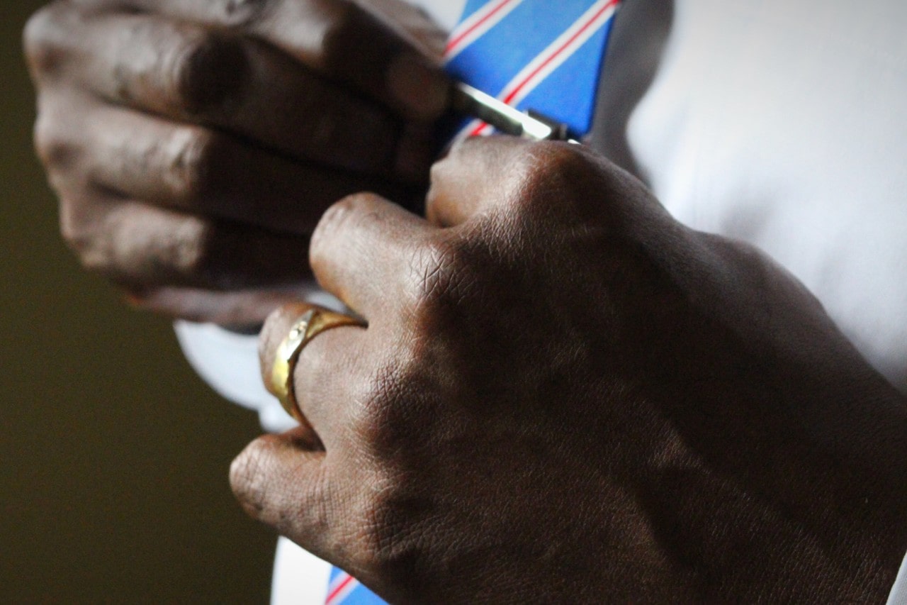 A close-up of a man adjusting a striped blue tie with a silver tie clip, wearing a gold wedding band on his ring finger.