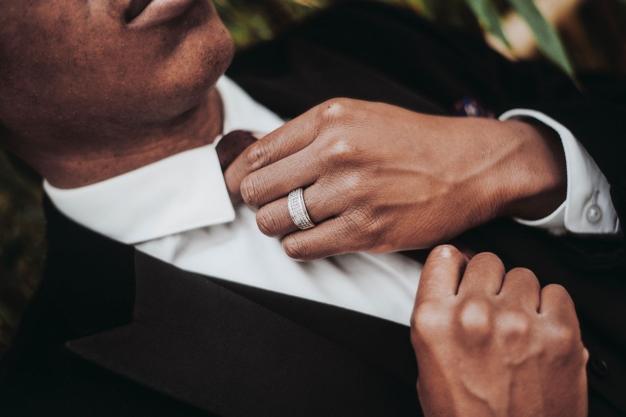 A groom in a suit adjusts his tie, showcasing a white gold diamond wedding band.