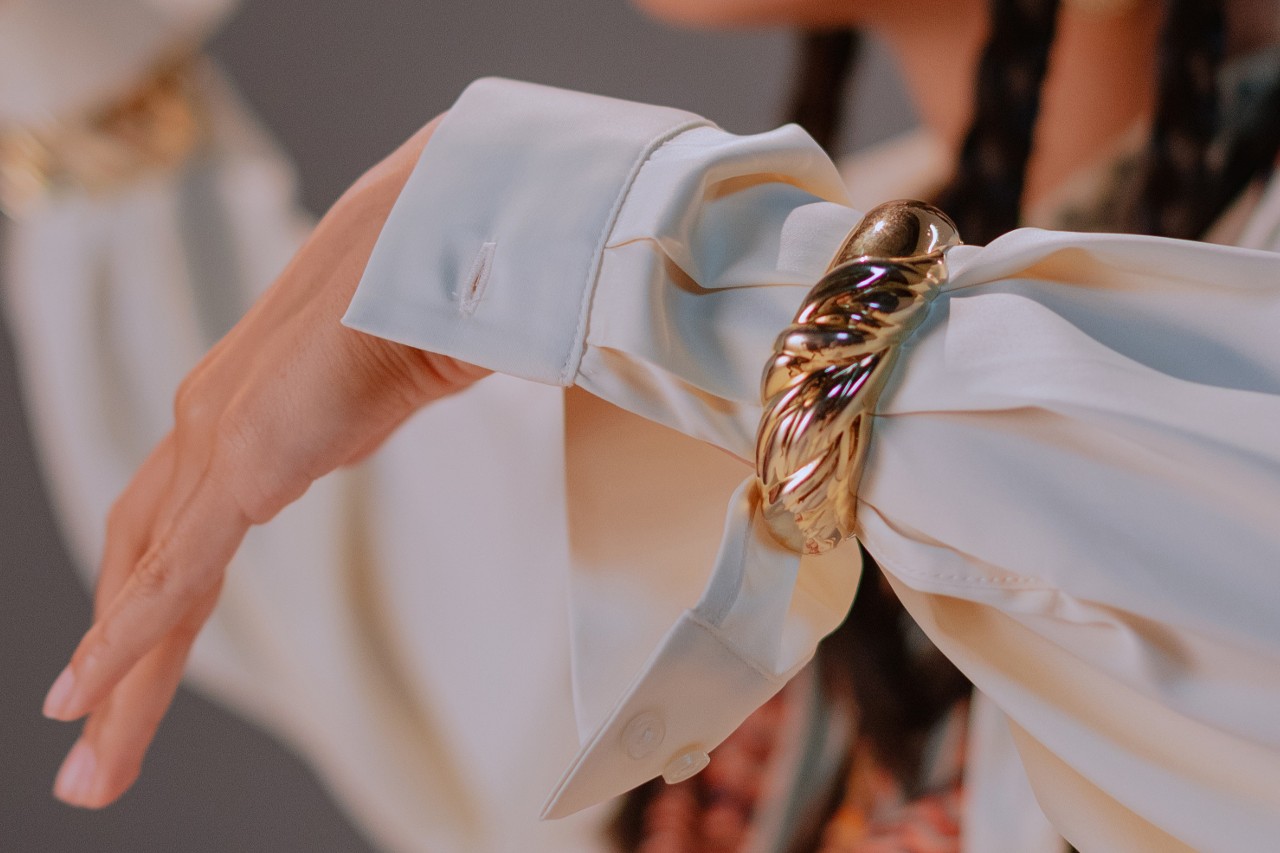 close up image of a woman’s wrist wearing a bold gold cuff bracelet over her shirt sleeve