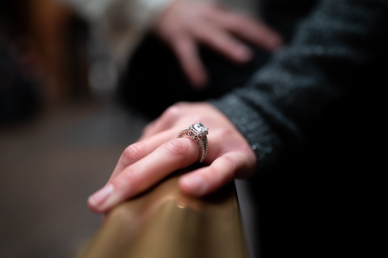 a woman’s hand on a banister, wearing an engagement ring, with her partner in the background
