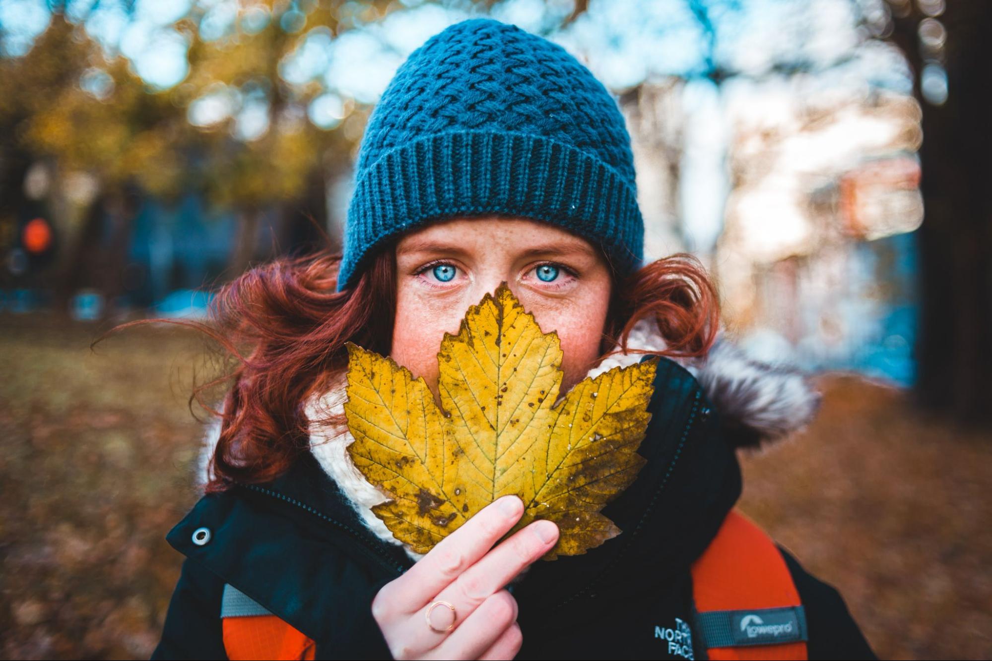 A redheaded woman concealing half of her face with a fallen leaf.