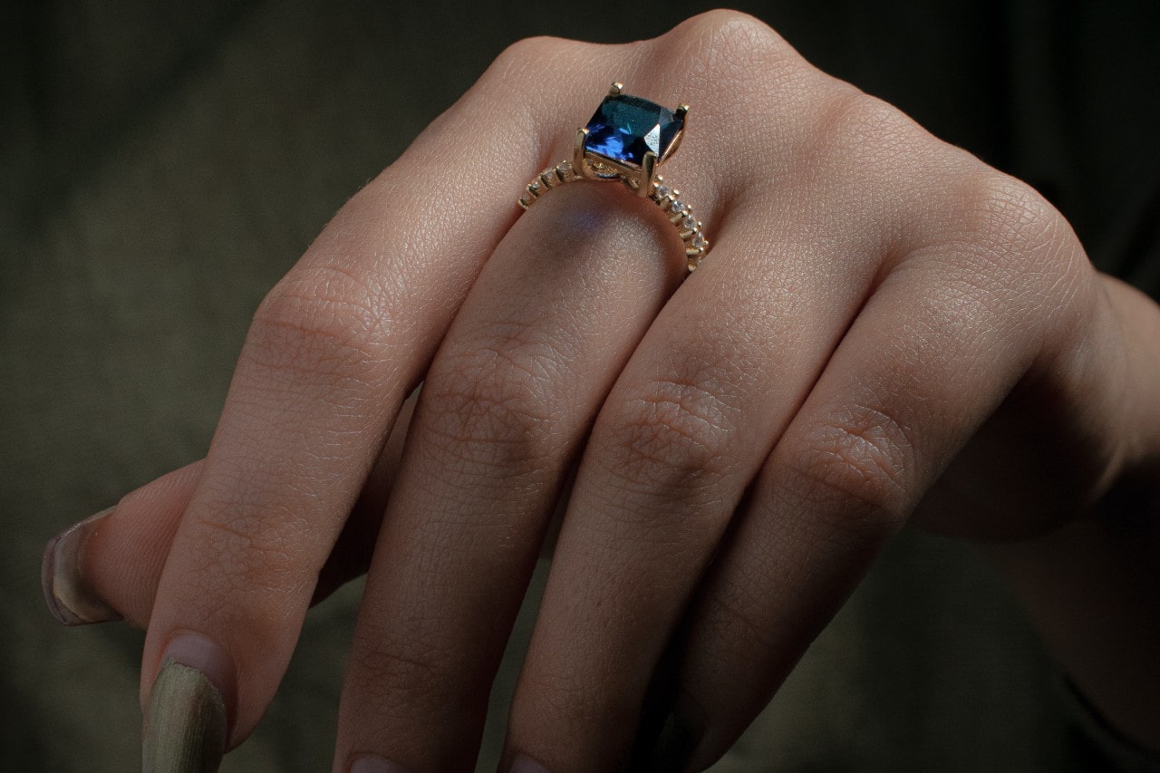 close up image of a hand wearing a yellow gold fashion ring featuring a princess cut sapphire