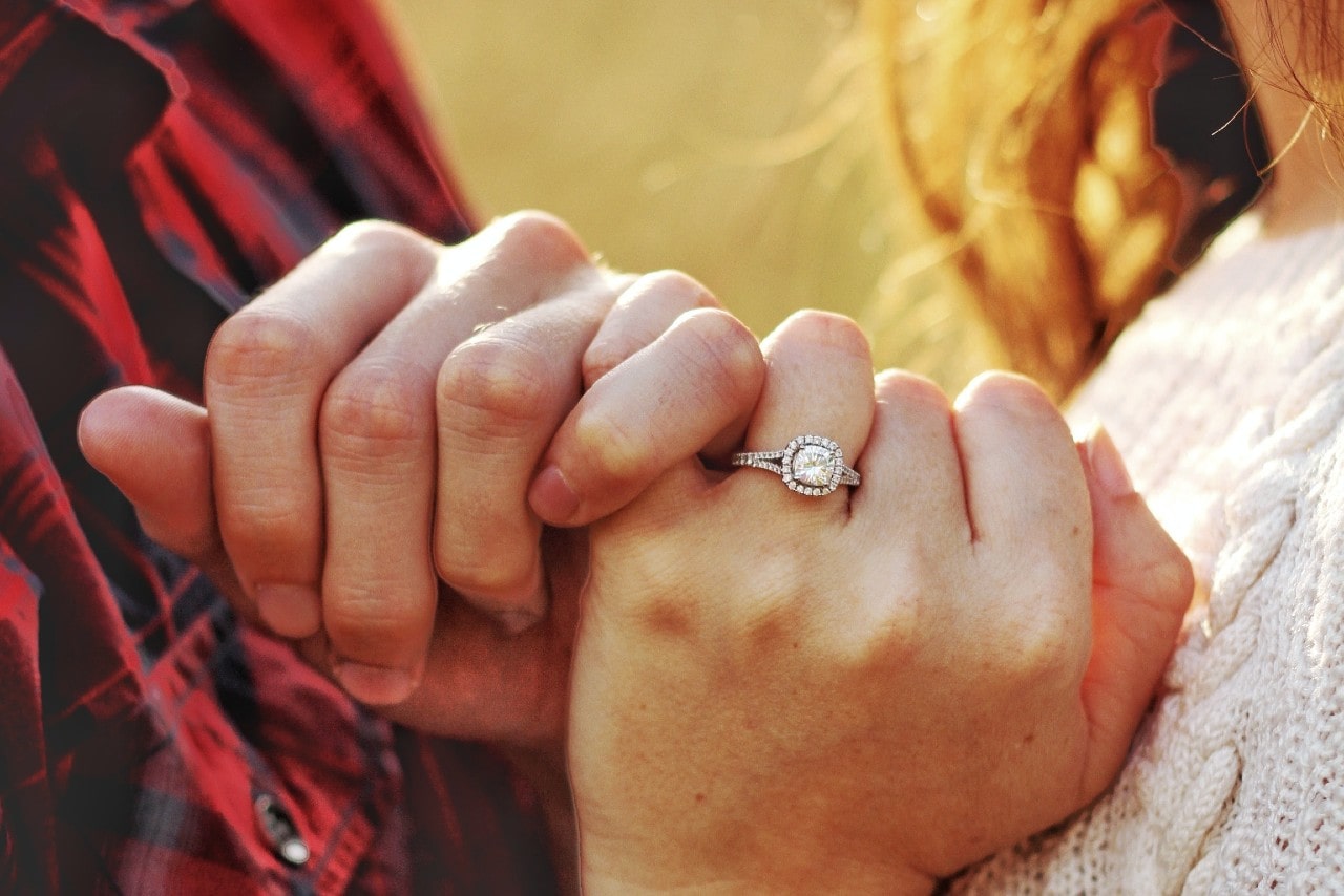 a couple intertwining their pinkies, the woman wearing a halo engagement ring