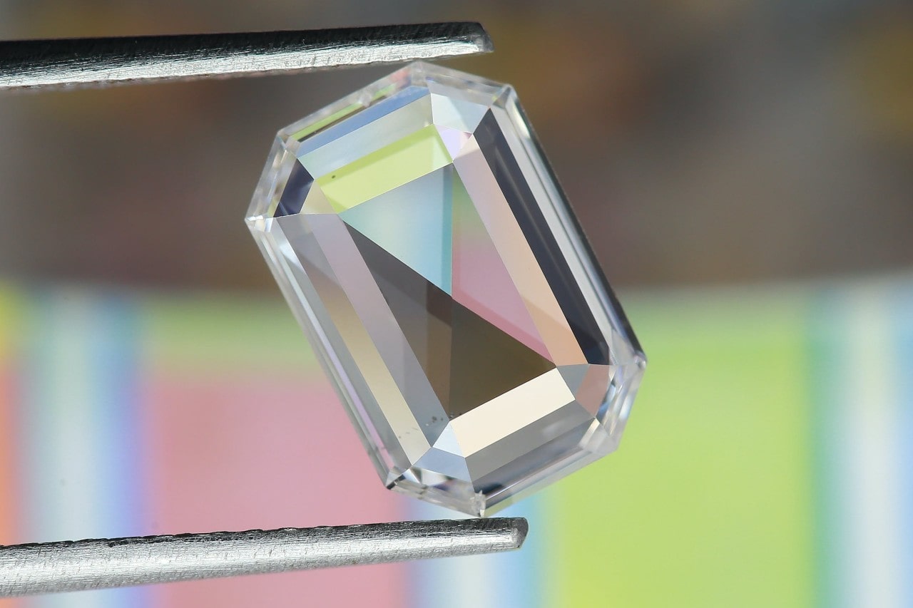 A close up of an emerald cut diamond held by tweezers against a blurry background