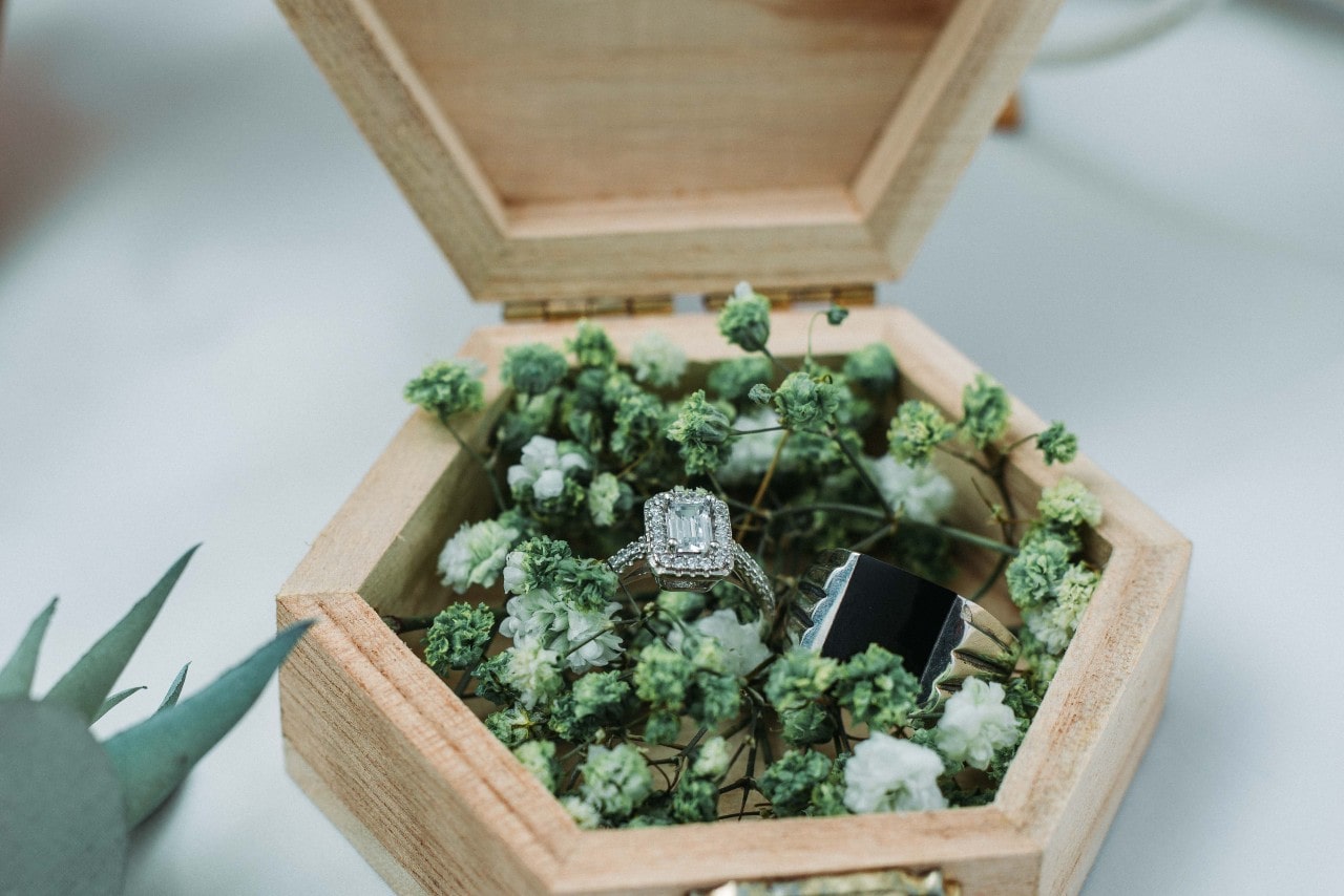 A white gold halo engagement ring and a black wedding band sit inside a wooden box filled with green and white flowers.