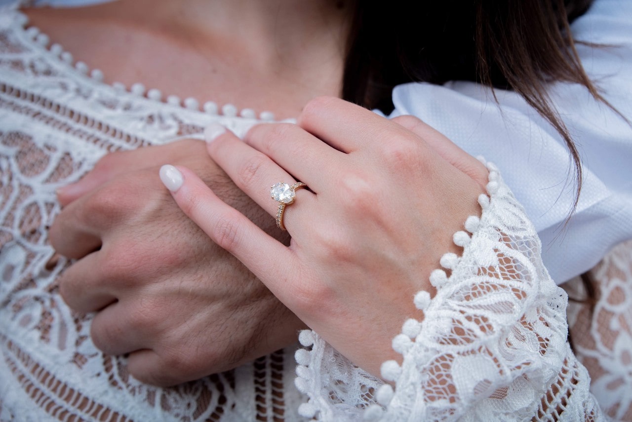 A woman's hand, adorned with an oval diamond engagement ring, rests on her partner’s hand.