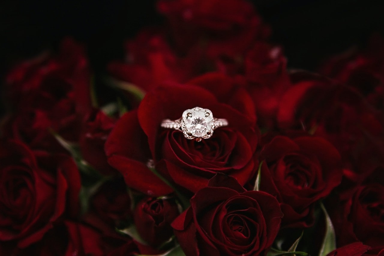 A rose gold side stone diamond engagement ring rests atop deep red roses.