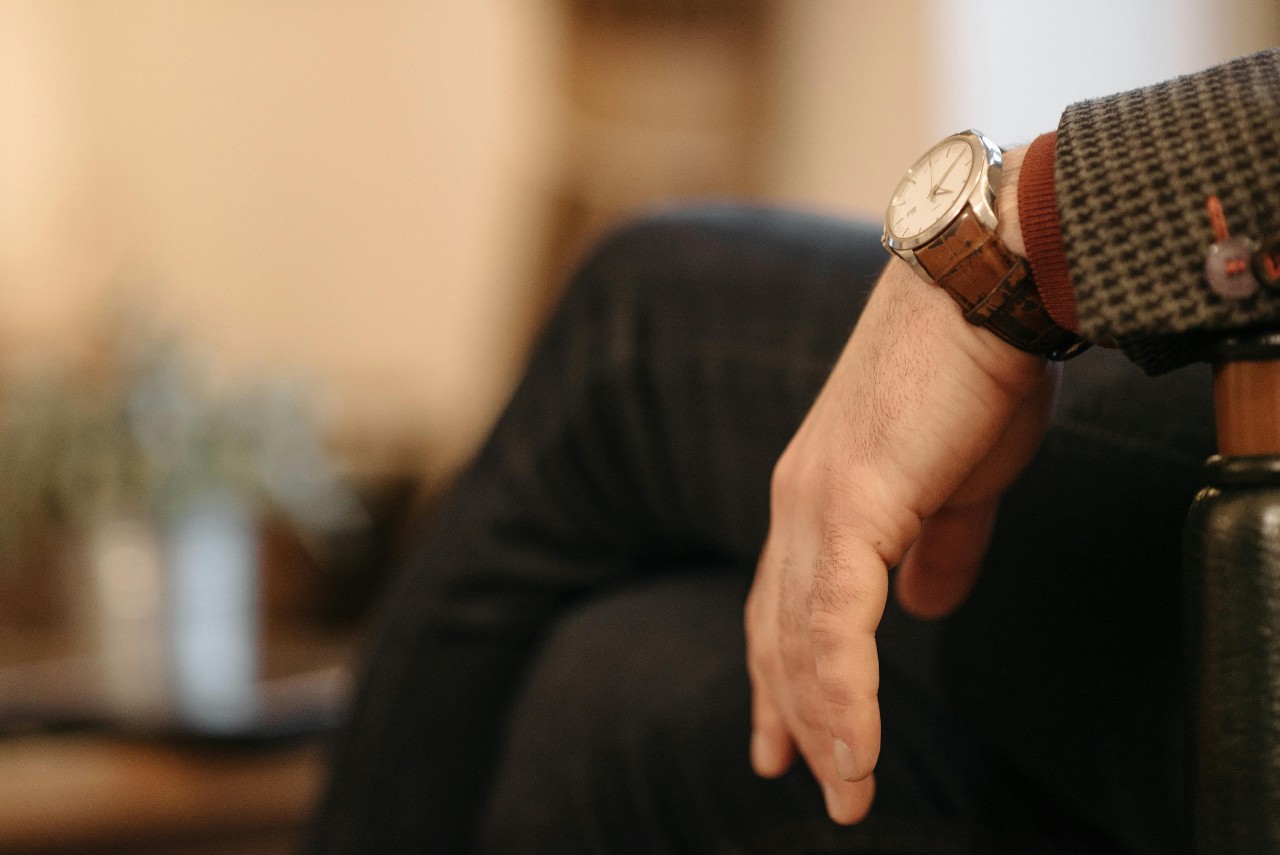A close-up of a man wearing a watch with a brown leather strap sitting in a chair and resting his arm on the armrest.