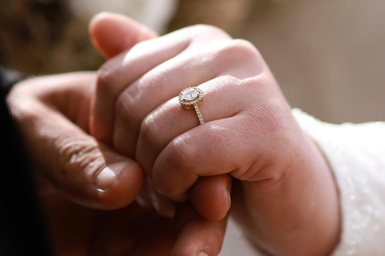 A close-up of a woman’s hand held in her partner’s hand, an elegant halo engagement ring on her finger.