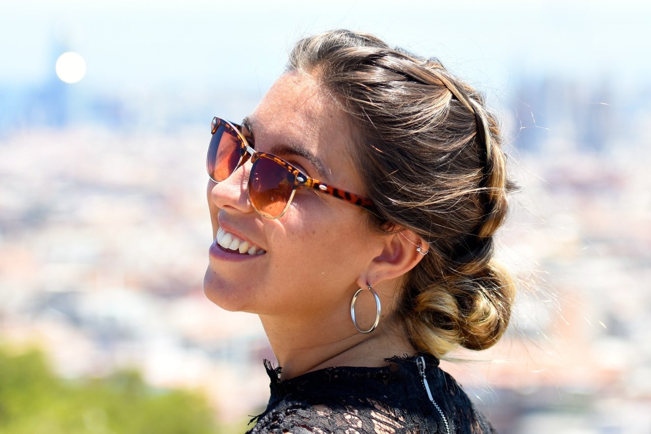 A woman smiling outdoors while wearing tortoiseshell sunglasses and large hoop earrings with her hair styled in a braid.