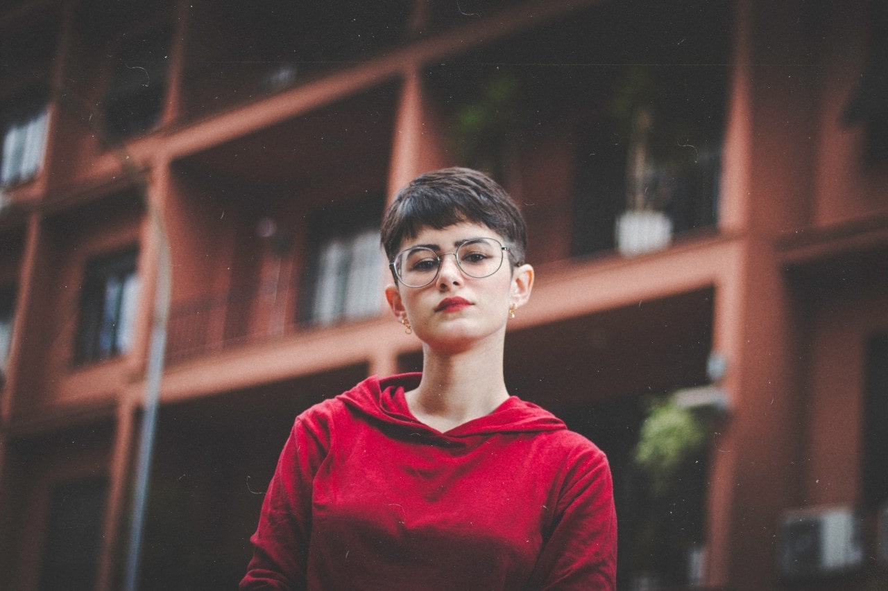 A woman wearing round glasses and small hoop earrings stands in front of an apartment complex.