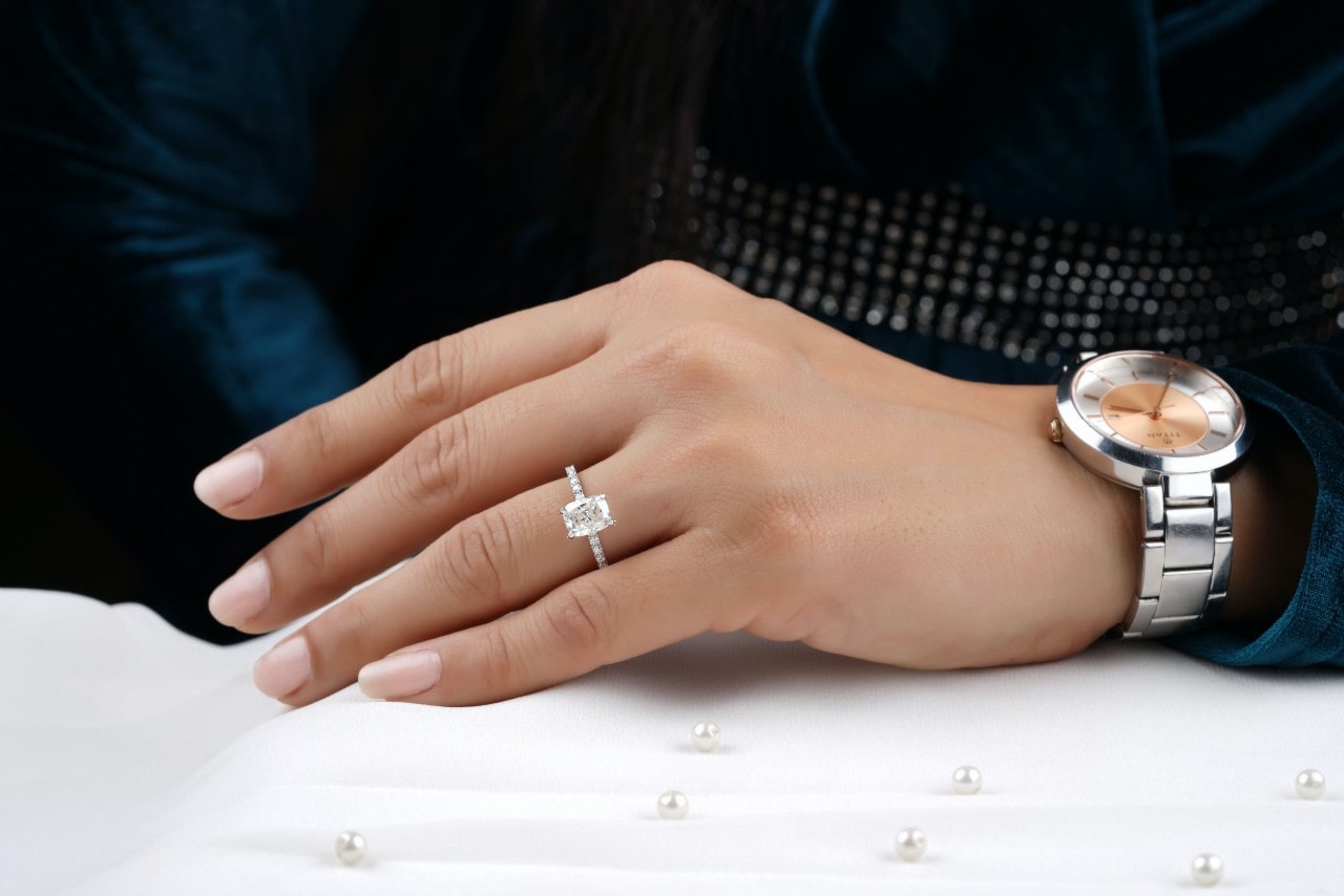 A woman's hand adorned with an emerald-cut diamond engagement ring and a silver wristwatch rests on a white surface decorated with scattered pearls. A woman's hand adorned with an emerald-cut diamond engagement ring and a silver wristwatch rests on a white surface decorated with scattered pearls.