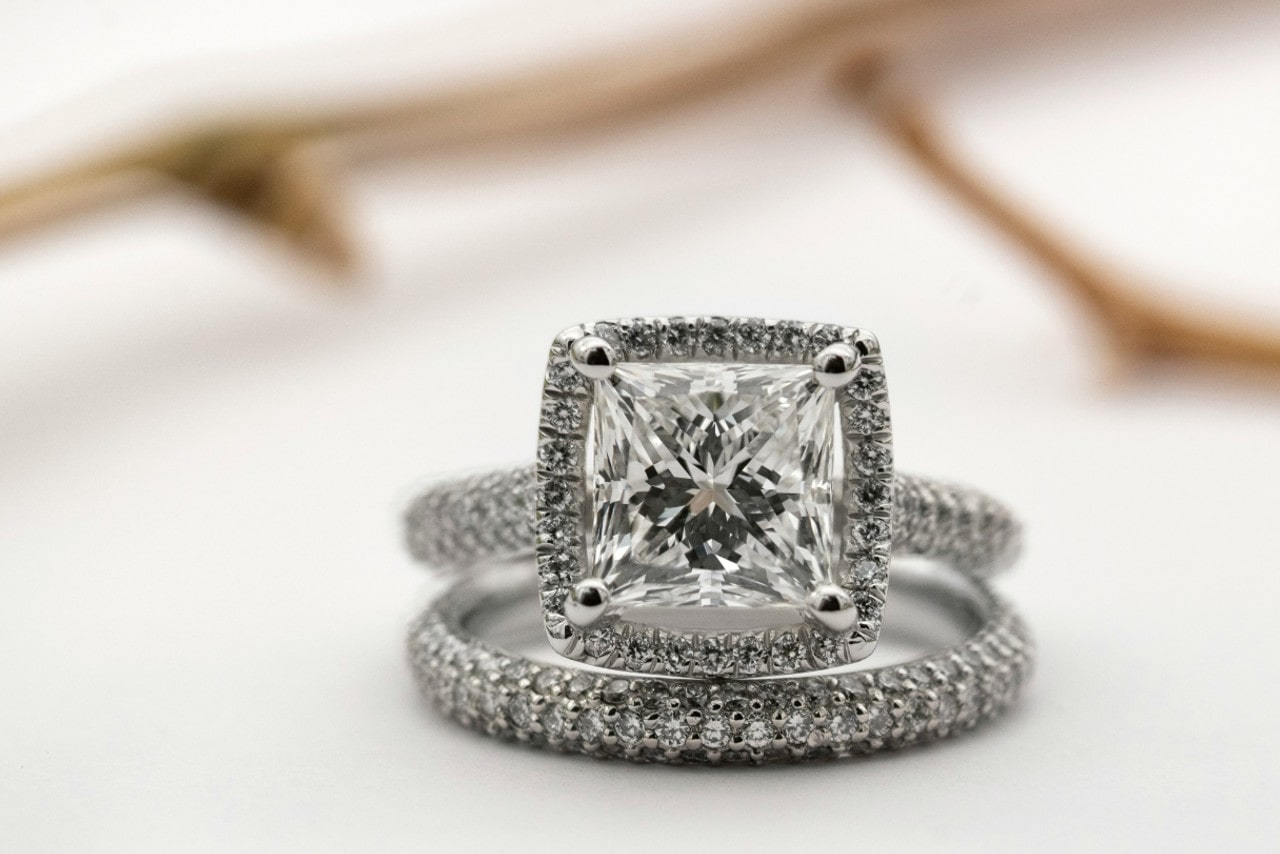 A close up of white gold princess-cut diamond engagement ring and wedding band against a blurry white background. A close up of white gold princess-cut diamond engagement ring and wedding band against a blurry white background.