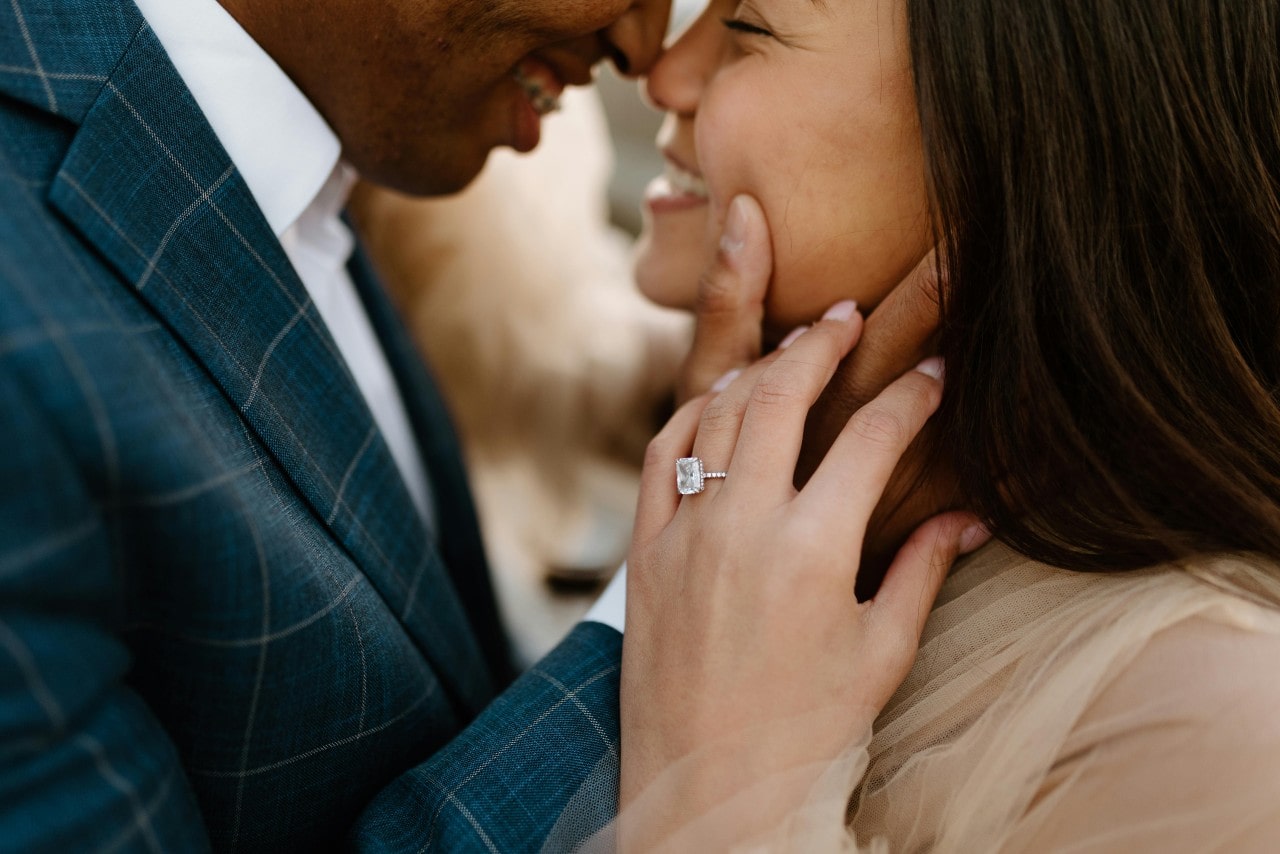 A couple smiling and leaning towards each other, showcasing a solitaire diamond engagement ring on a woman’s hand. A couple smiling and leaning towards each other, showcasing a solitaire diamond engagement ring on a woman’s hand.