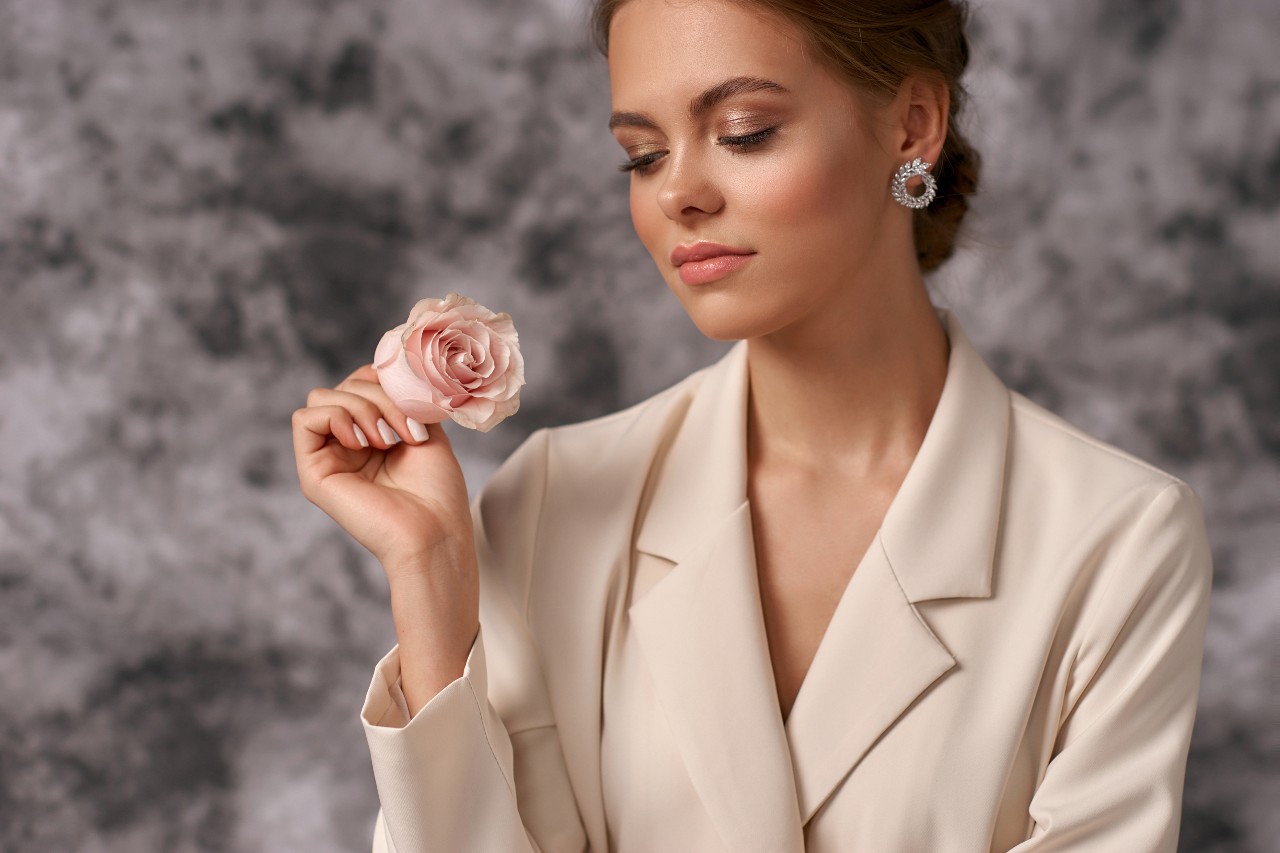 A woman in a cream blazer holds a pink rose, wearing diamond earrings against a softly textured gray background.