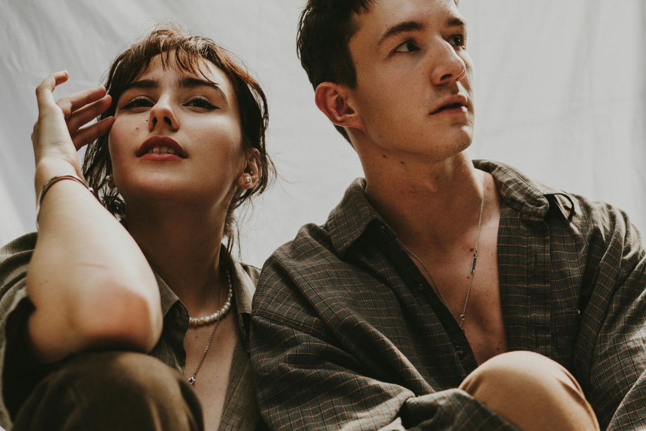 A woman wearing pearl earrings, a delicate bracelet, and a pearl necklace sits beside a man in a checkered shirt with a minimalist chain necklace, both styled in neutral-toned attire.