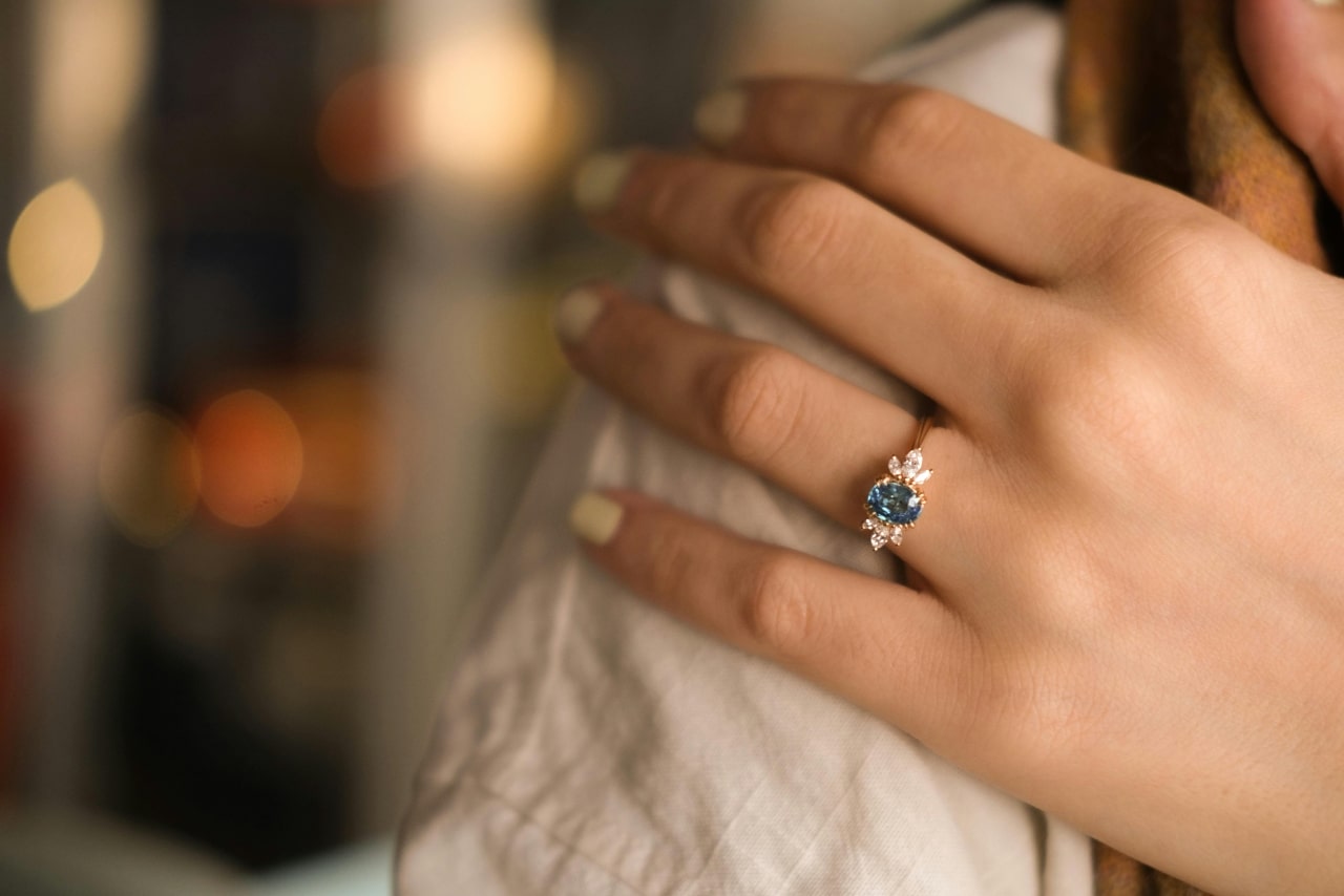 A close-up of a hand with pale nail polish on the shoulder, adorned with an elegant three-stone engagement ring featuring a blue center gemstone flanked by two side diamonds.