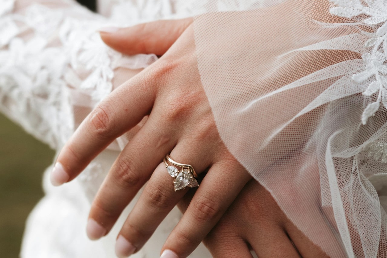 Close-up of a bride’s crossed hands, showcasing an elegant three-stone gold engagement ring over a white lace wedding dress.