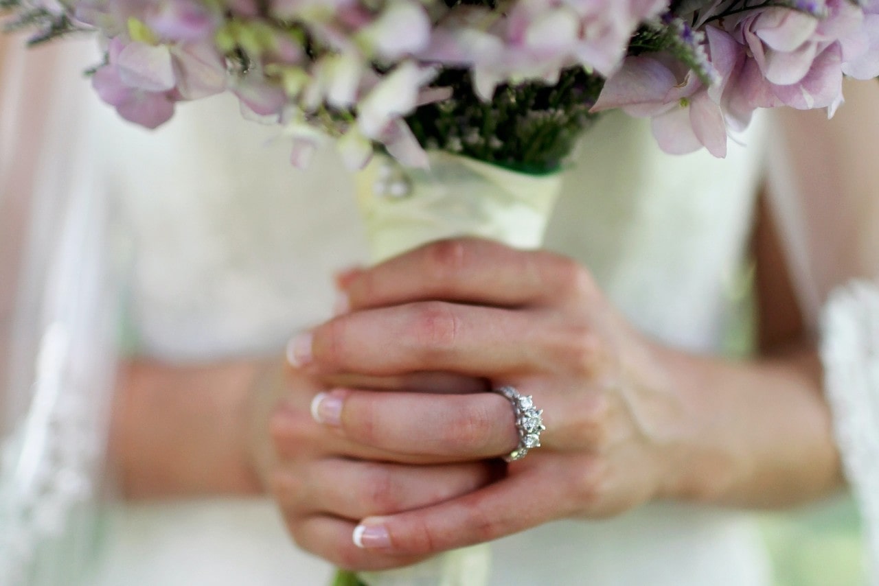 Close-up of a bride’s hands holding a bouquet of light purple flowers, showcasing a sparkling three-stone diamond ring.