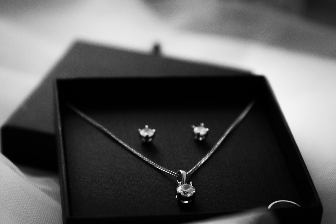 A black gift box displays a matching silver jewelry set featuring a diamond solitaire pendant necklace and a pair of diamond stud earrings, arranged against a dark fabric background.