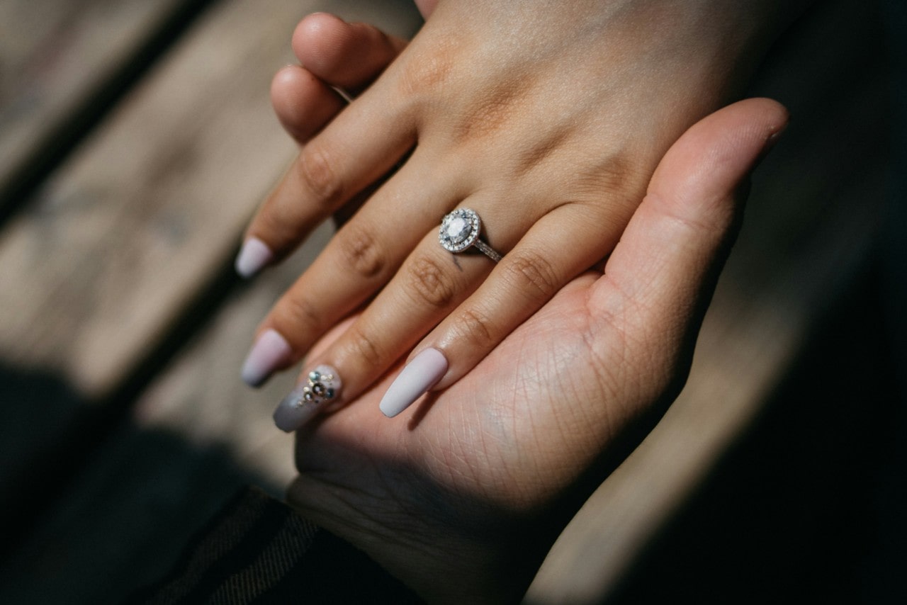 A woman’s hand on top of the man’s hand, showcasing long nail and diamond white gold halo engagement ring A woman’s hand on top of the man’s hand, showcasing long nail and diamond white gold halo engagement ring