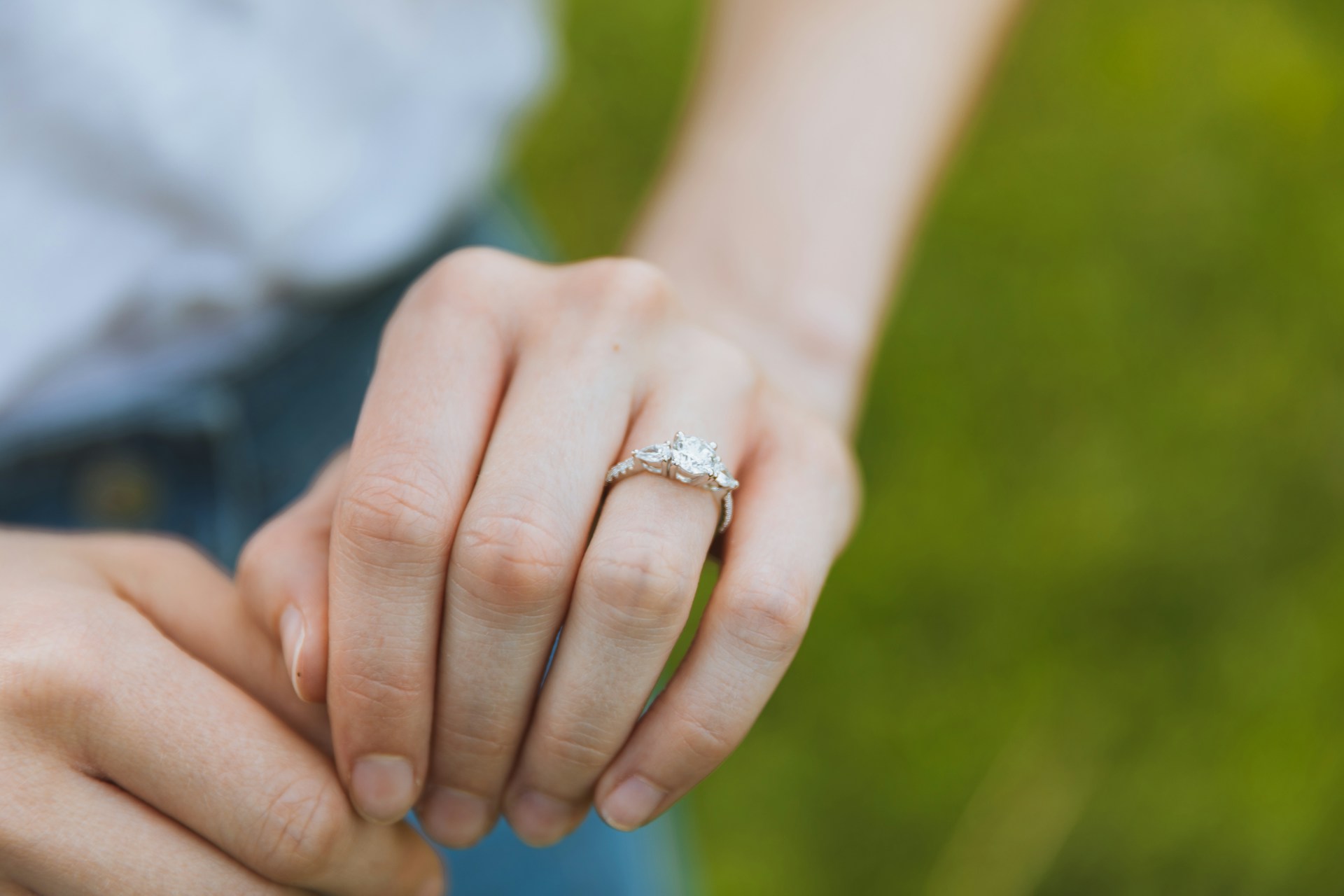 A close up of a three stone engagement ring on a woman’s hand against a blurry grass A close up of a three stone engagement ring on a woman’s hand against a blurry grass