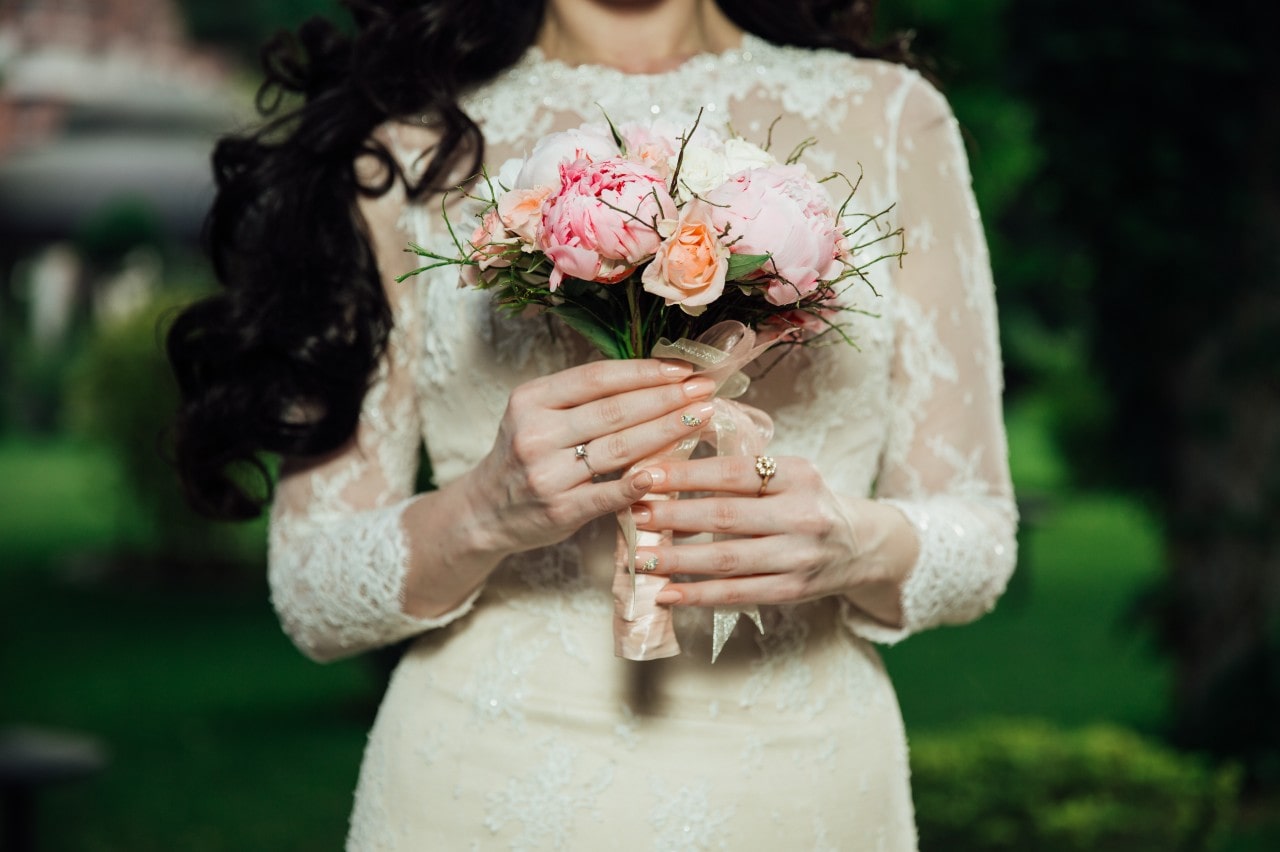 A bride in a lace wedding dress holds a bouquet of pink and peach flowers tied with a ribbon, showcasing her yellow gold halo engagement ring A bride in a lace wedding dress holds a bouquet of pink and peach flowers tied with a ribbon, showcasing her yellow gold halo engagement ring