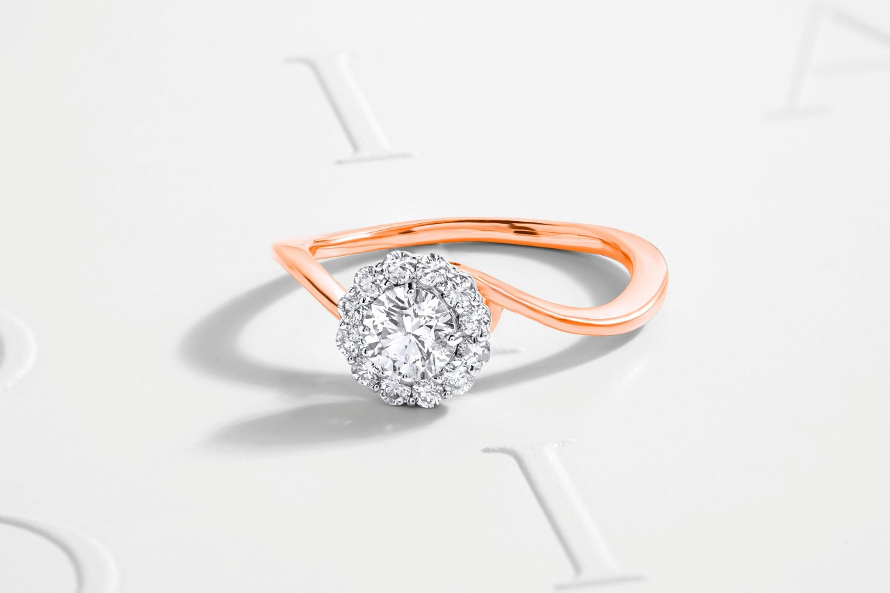 A close-up of a mixed metal engagement ring with a curving band on a white surface. A close-up of a mixed metal engagement ring with a curving band on a white surface.